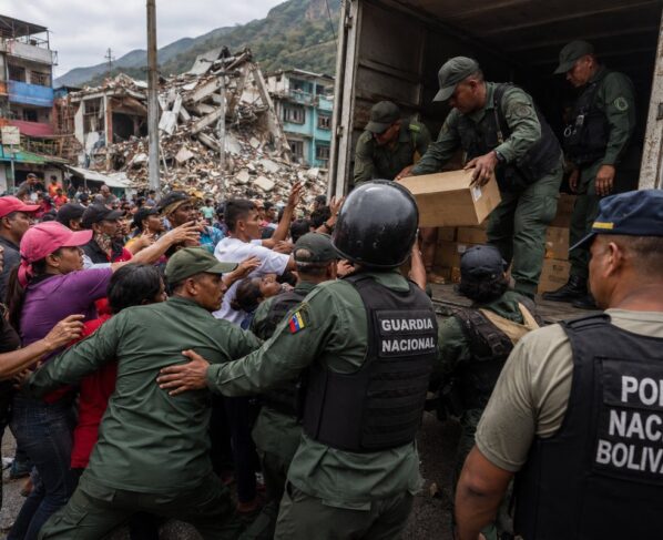 Editorial image showing security forces controlling humanitarian supplies before a crowd of civilians in an earthquake-affected urban area, used to illustrate allegations of aid diversion and obstacles to rescue efforts in Venezuela.